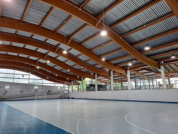 Vista de la pista polideportiva, con portería y canasta al fondo y una cubierta ondulada de vigas de madera