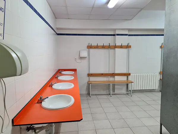 Vista de los aseos de los vestuarios, con una fila de lavabos y un secamanos y bancos de madera con perchas al fondo
