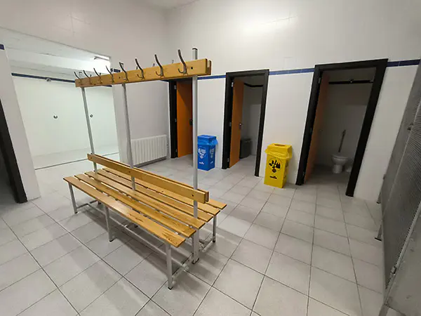 Vestuario con un banco, perchas, aseos al fondo y área de duchas a la izquierda