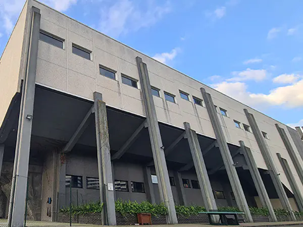 Vista lateral del exterior del edificio, con plantas ornamentales en un pasillo bajo sus columnas y una mesa de ping-pong