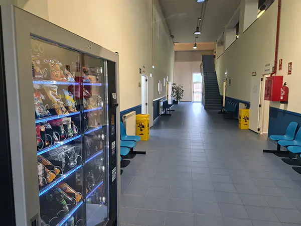 Pasillo interior, con sillas, una máquina expendedora y, al fondo, una escalera de acceso a la planta superior