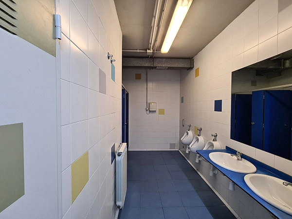 Pasillo de los aseos, con lavabos, urinarios y un espejo a la derecha