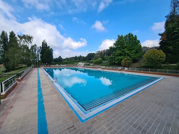 Panorámica de de la piscina principal, con las vallas perimetrales y rodeada de jardines y árboles