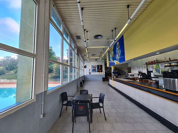 Interior de la cafetería, con grandes ventanales con vistas a la piscina, mesas, sillas y barra de servicio a la derecha