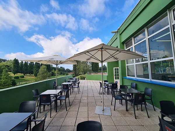 Exterior de la cafetería, con mesas, sillas y sombrillas. Vistas de la piscina principal y el Parque de Invierno