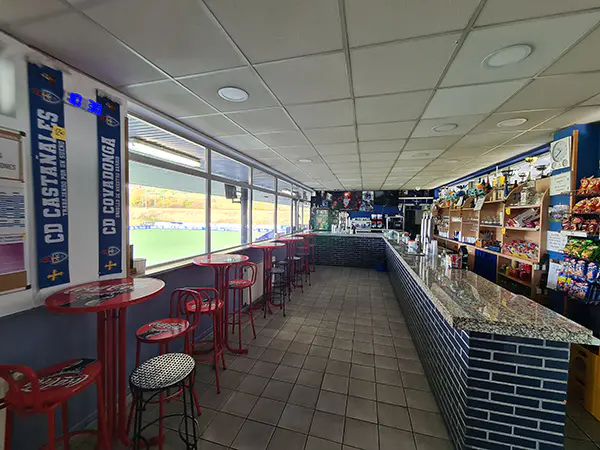 Cafetería del estadio. Amplio ventanal lateral vistas campo de juego. Interior sillas, mesas en rojo. Barra del bar en L