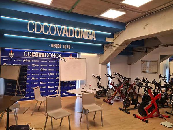 Sala técnica con sillas, mesa y pizarra delante muro con logos de diferentes marcas en letras blancas. Bicicletas estáticas