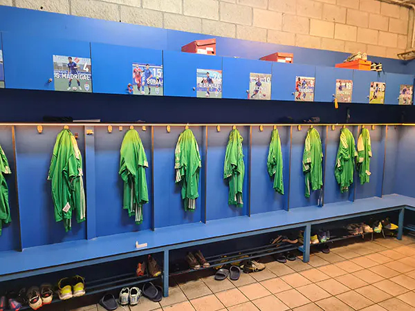 La equipación verde del equipo de fútbol está colgada bajo la foto de cada jugador, sobre los bancos zapateros azules