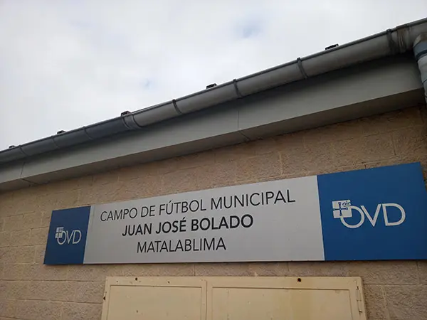 Cartel azul a ambos lados con el logo de Oviedo en letra blanca. Sobre fondo blanco: Campo de Fútbol,  Juan José Bolado
