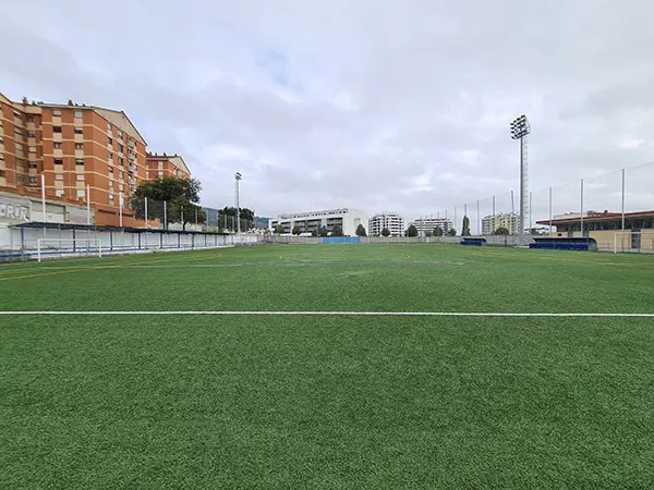 Vista del césped artificial campo de fútbol, destacan la portería a la izquierda, dos torres de iluminación y bancos equipos