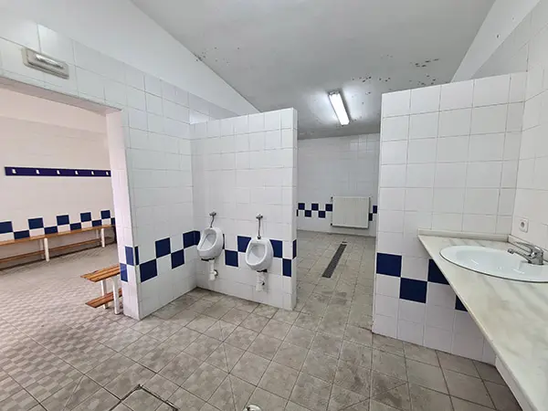 Zona de aseos del vestuario con azulejos blancos y dos hileras de alternos azules y blancos. Además, dos urinarios y lavabo
