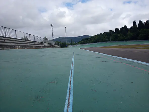 Vista a pie de pista del velódromo y las y gradas laterales, con torres de iluminación y vegetación al fondo
