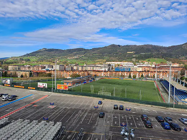 Vista aérea del campo de fútbol Hermanos Llana, con el aparcamiento delante, edificios alrededor y montañas al fondo