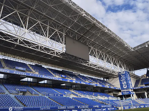Tribuna lateral con filas de gradas, vallas publicitarias a pie de campo, el techo metálico y el marcador