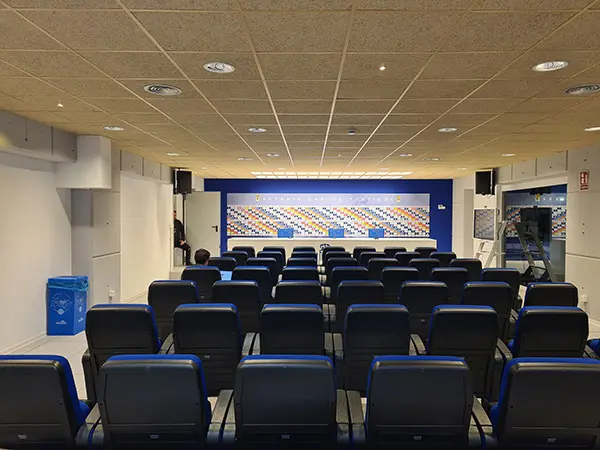 Sala de prensa con filas de asientos azules y al fondo mesa para conferenciantes, detrás logos de diferentes empresas
