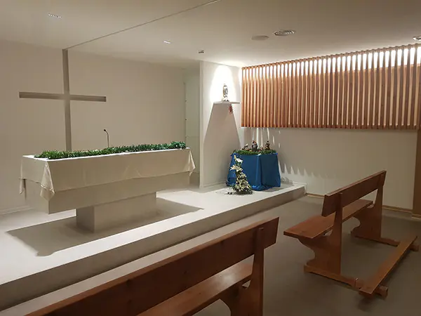 Interior de la capilla con bancos de madera, altar con decoración navideña, detrás una gran cruz esculpida en la pared 