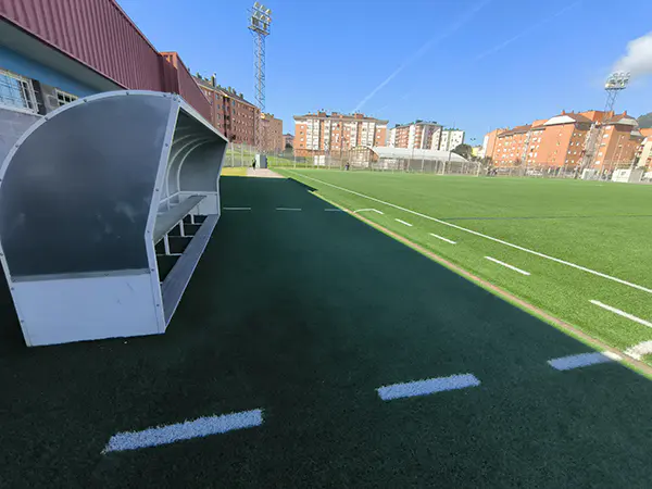 Vista lateral a pie de campo del terreno de juego, con los banquillos en primer plano y edificios al fondo