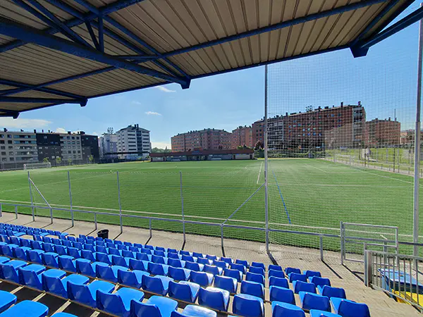 Vista lateral del terreno de juego desde las gradas, con las vallas de protección y edificios alrededor