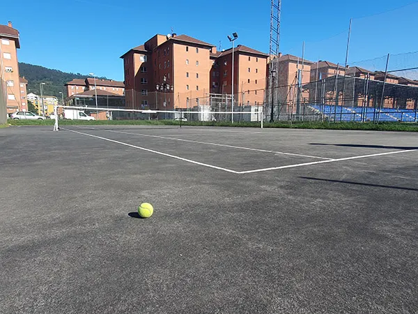 Vista lateral de una de las pistas de tenis, con una pelota en primer término y edificios de viviendas al fondo