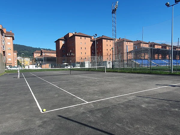 Vista lateral de una de las pistas de tenis, con una pelota en primer término y edificios de viviendas al fondo