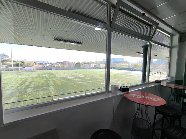 Campo de fútbol a través de los ventanales de la cafetería con mesas altas rojas con taburetes