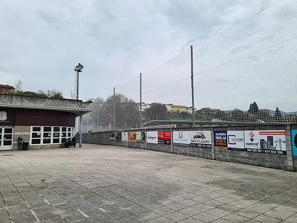 Muro exterior del campo de fútbol y red de protección blanca para evitar la salida de balones. A la izquierda la cafetería