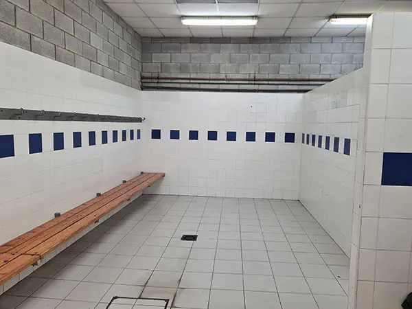 Interior del vestuario alicatado en blanco intercalado con una hilera de azulejos en azul con bancos y percheros de madera