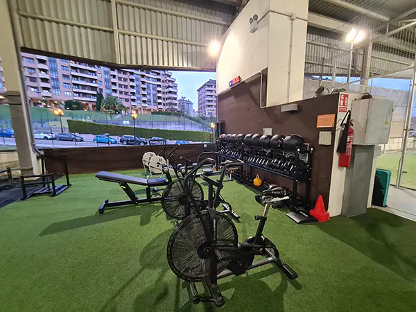 Pequeña zona exterior de ejercicio cubierta, con bicicletas y bancos de musculación. Vista de edificios al fondo