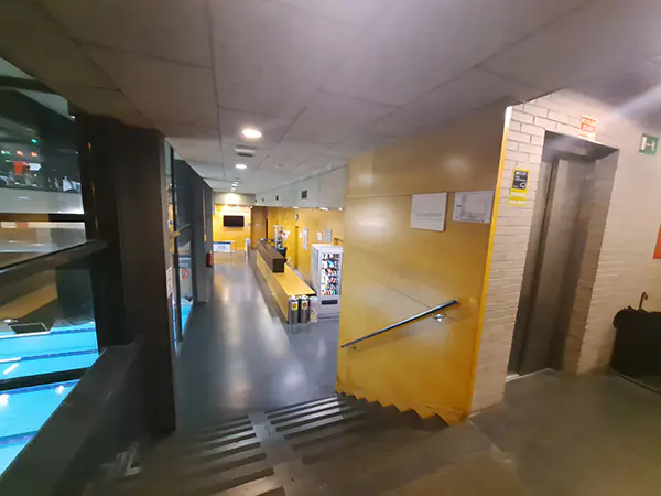 Entrada al recinto del centro deportivo, con escaleras de acceso y ascensor en primer plano y mostrador de recepción al fondo