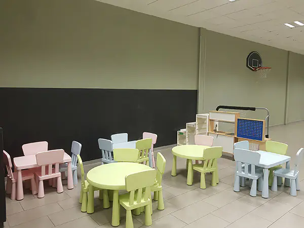 Área de servicios infantiles, con unas pequeñas sillas y mesas de diferentes colores y una canasta de minibasket