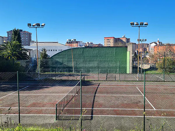 Vista lateral de una pista de tenis, con la red central, vallas, torres de iluminación y, al fondo, la pared de un frontón