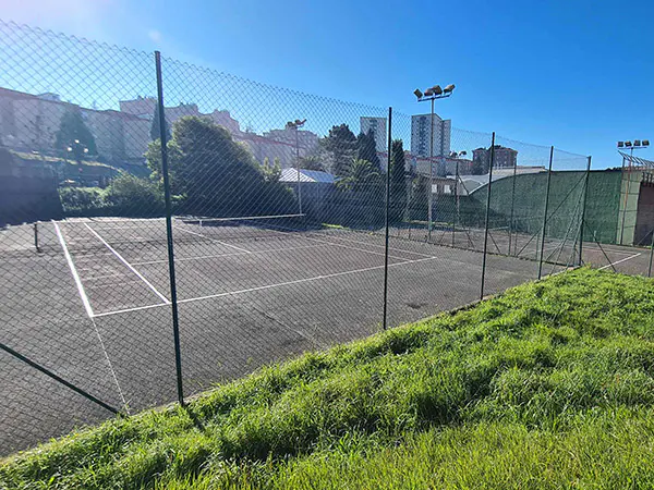 Vista frontal de una pista de tenis, desde el exterior de la valla perimetral, con edificios circundantes al fondo