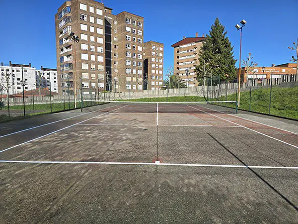 Vista frontal de una de las pistas de tenis, con el muro de cierre de las instalaciones y edificios circundantes al fondo
